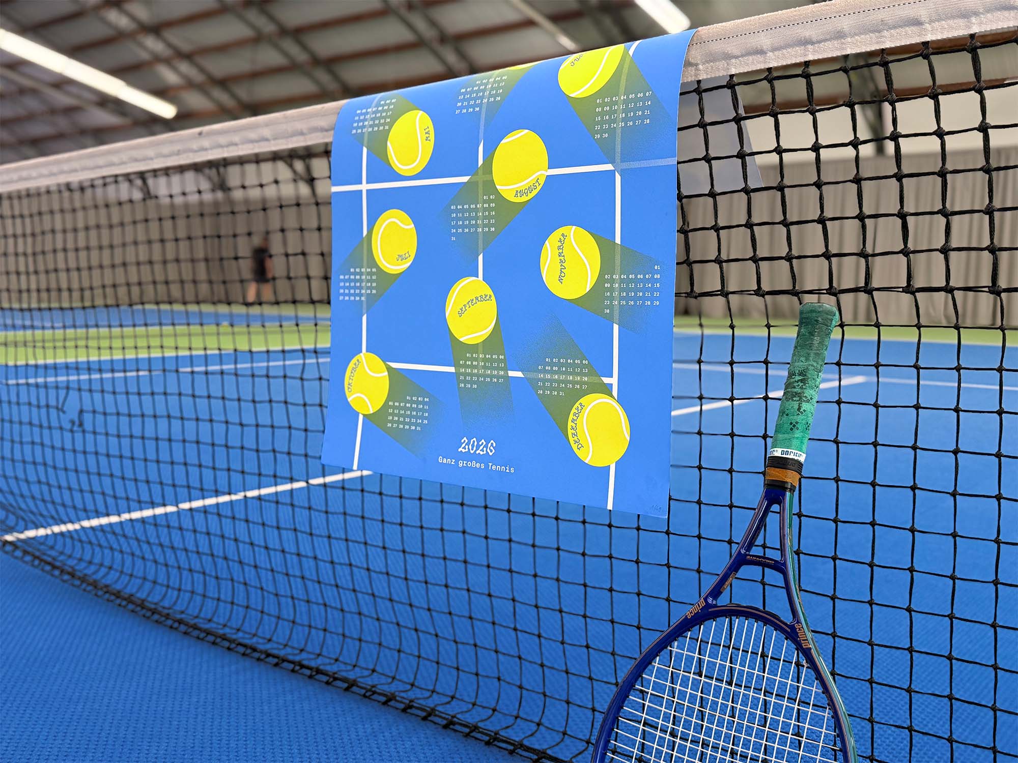 Photo of our 2026 studio_upstruct calendar draped over a tennis net on a blue tennis court, with a tennis racket leaning against the net.