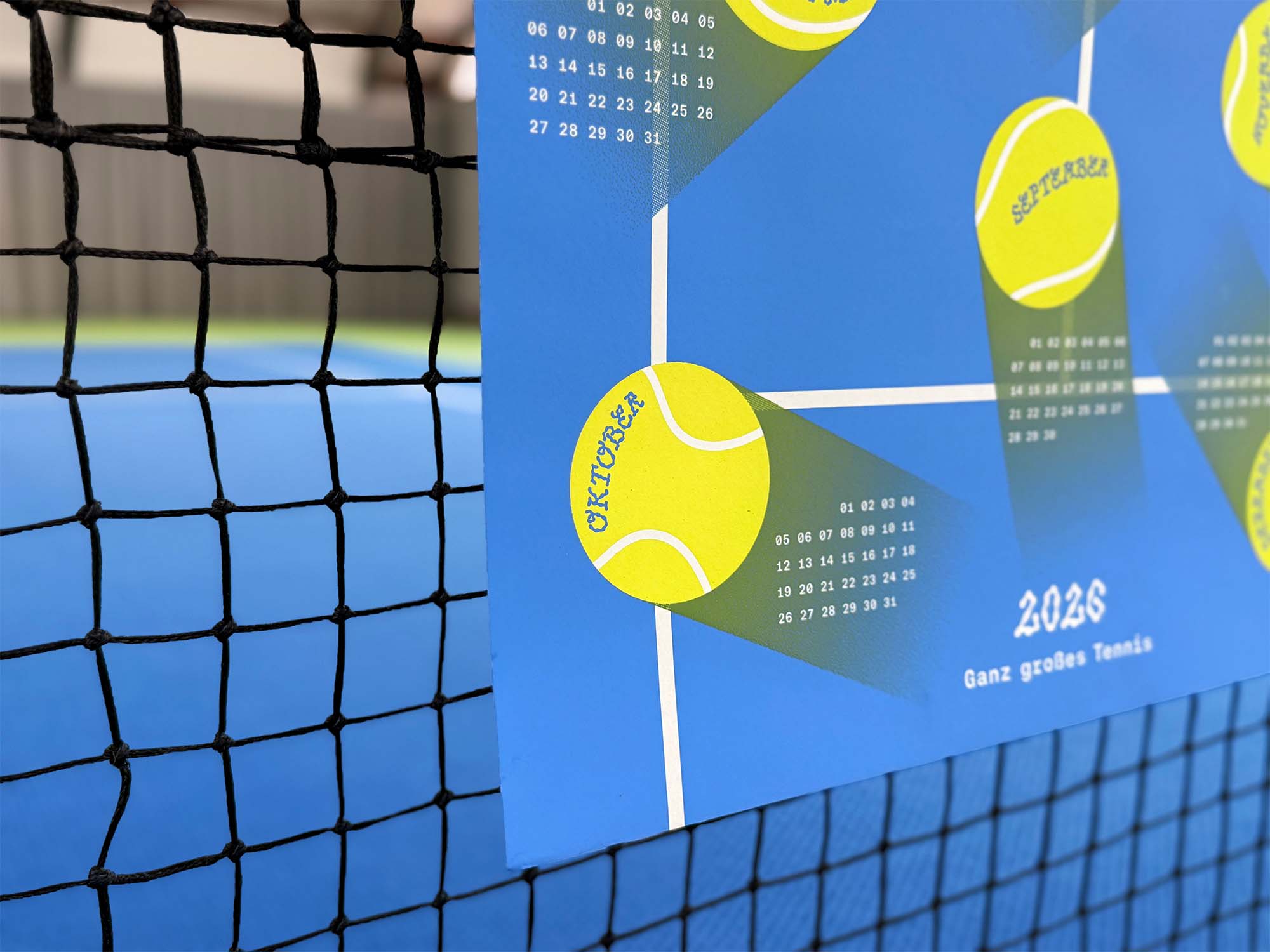 Close-up of the studio_upstruct calendar draped over a tennis net on a blue tennis court, showing a few months and the calendar title, '2026 – Ganz großes Tennis.'