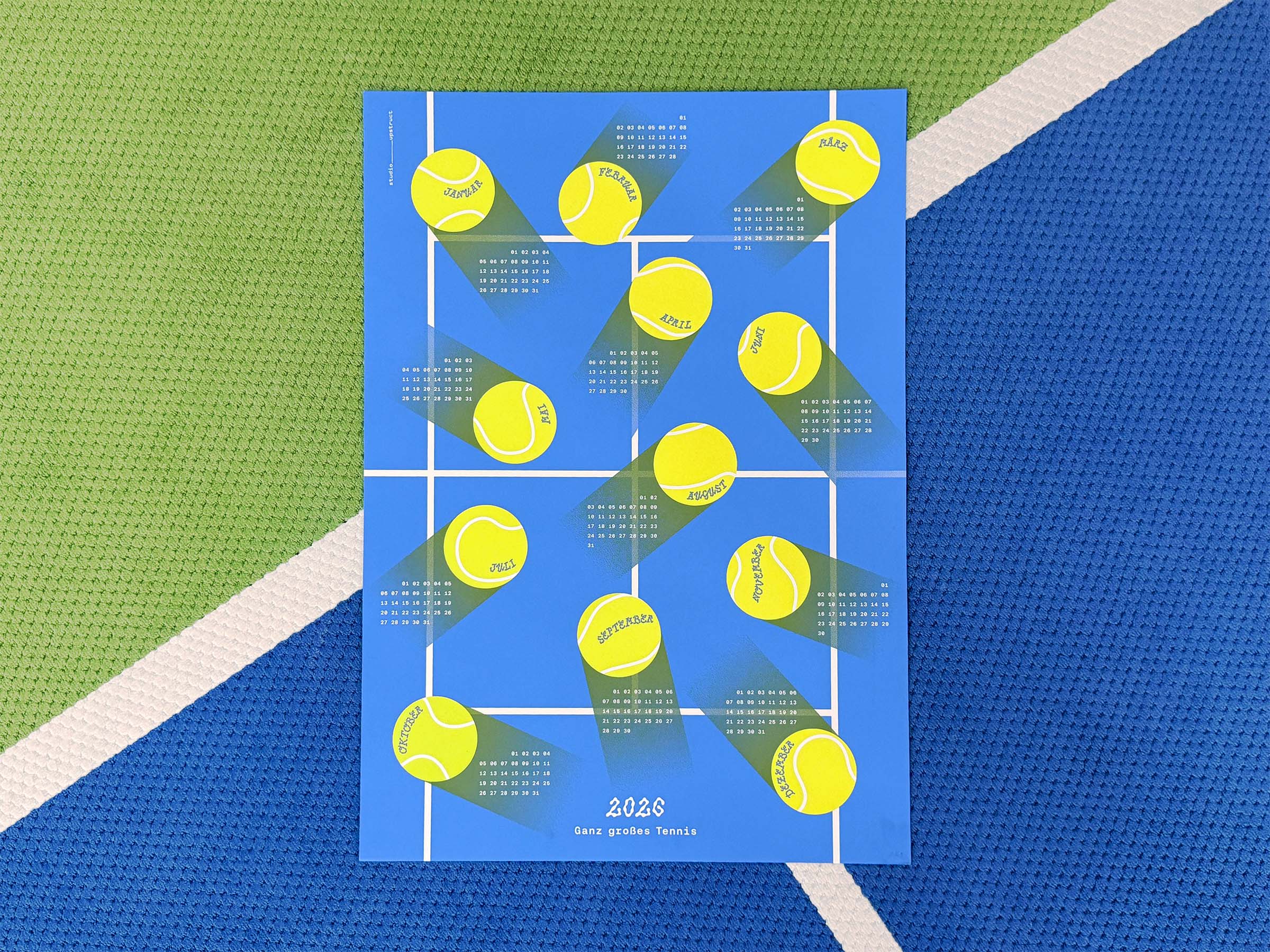 Full view of the 2026 studio_upstruct calendar, '2026 – Ganz großes Tennis.' The calendar is laid out like a blue tennis court, with 12 large yellow balls showing each month. Each ball leaves a trail behind it, showing the calendar dates of the respective month.